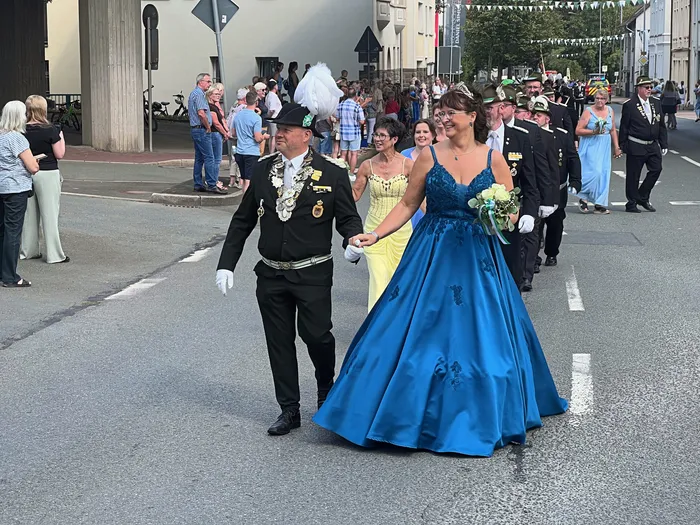 Ein Mann in schwarzer Schützenuniform mit Federhut und eine Frau in einem großen, blauen Ballkleid gehen Hand in Hand bei einem Festumzug.