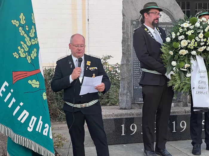 Ein Mann in Schützenuniform spricht in ein Mikrofon vor einem Ehrenmal, während ein Mann daneben einen großen Blumenkranz hält.