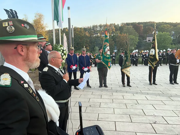 Ein Mann in Uniform spricht bei einer Gedenkfeier des Schützenfestes Eiringhausen, während Männer im Hintergrund zeremonielle Fahnen halten.