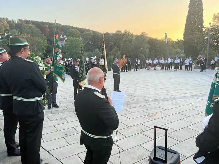 Männer in Schützenuniformen halten Fahnen bei einer Gedenkfeier auf einem gepflasterten Platz im Abendlicht. Ein Mann liest aus einem Dokument vor.