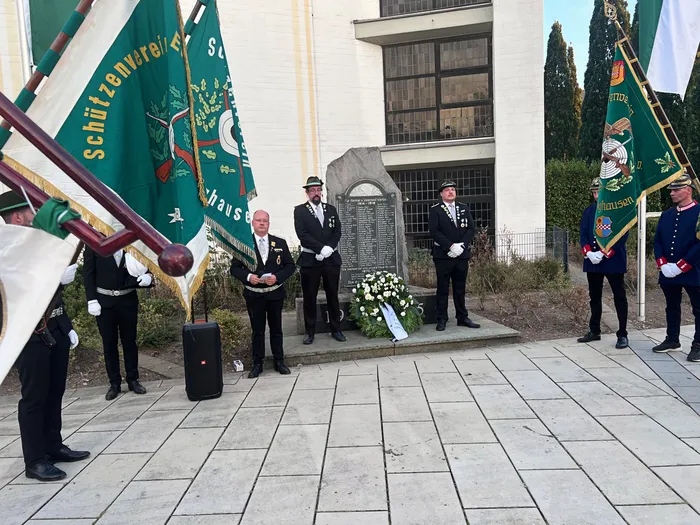 Männer in Schützenuniformen stehen bei einer Gedenkfeier an einem mit einem weißen Blumenkranz geschmückten Ehrenmal.