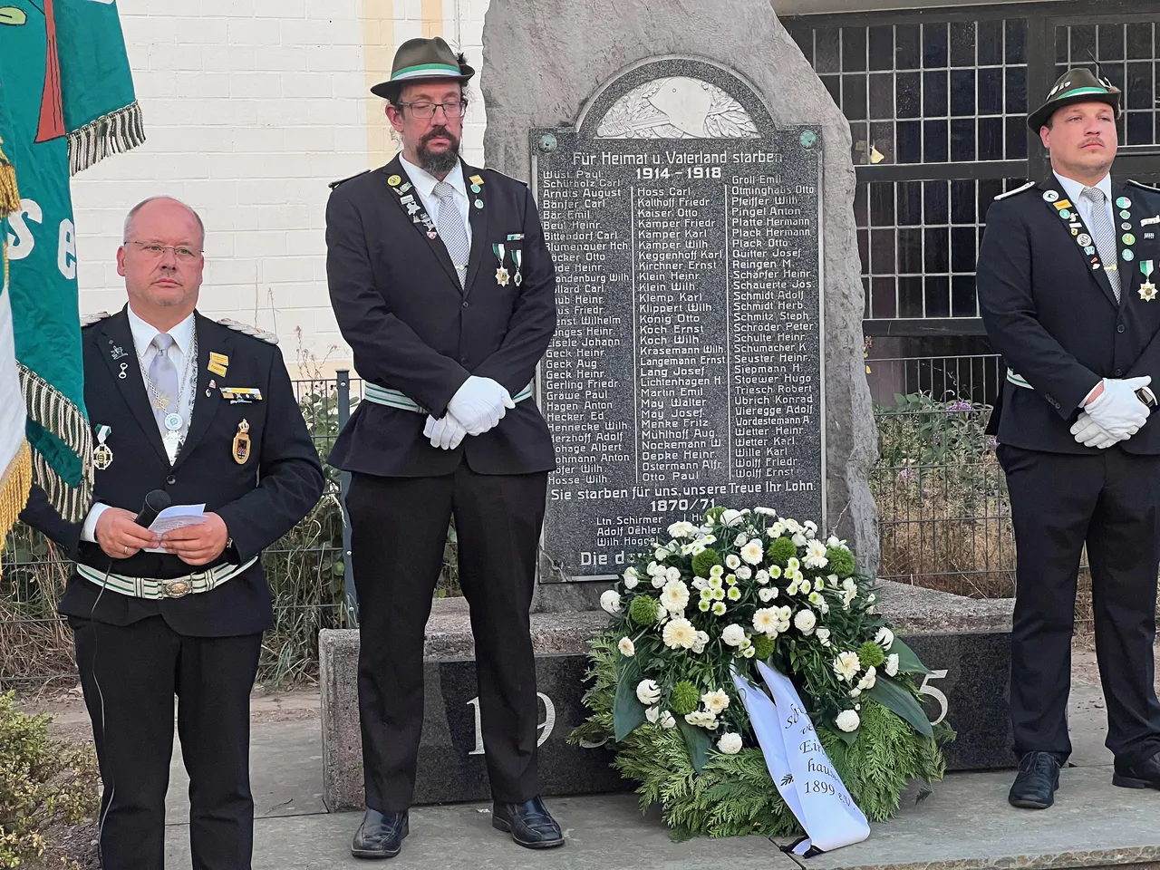 Drei Männer in traditionellen Schützenuniformen stehen vor einem steinernen Ehrenmal, vor dem ein Blumenkranz liegt.
