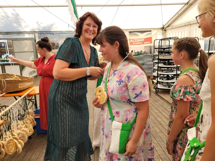 Eine Frau in einem grünen Kleid lächelt ein Mädchen an, das ein Gebäckstück hält. Im Hintergrund stehen Körbe mit Gebäck auf einem Tisch.