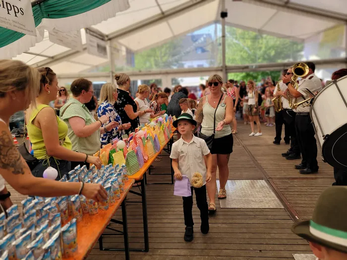 Ein Junge mit Hut steht vor einem Tisch mit bunten Tüten beim Kids Day des Schützenfestes Eiringhausen. Im Hintergrund spielt eine Blaskapelle.