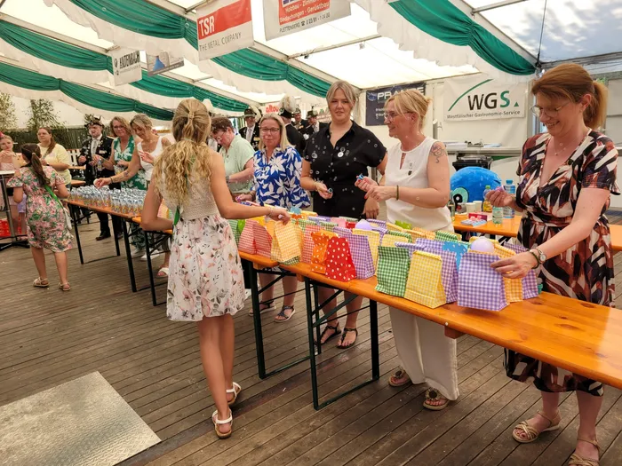 Frauen stehen hinter einem langen Holztisch mit zahlreichen bunten, gemusterten Geschenktüten unter einem Festzelt.