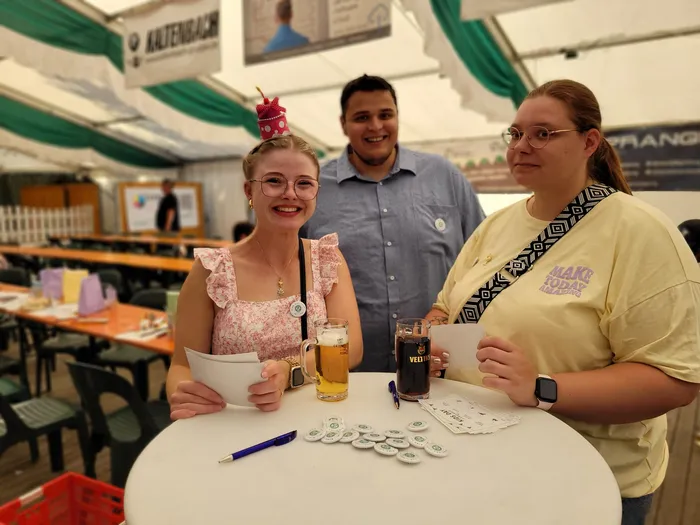 Drei lächelnde Personen stehen hinter einem Tisch mit Getränken und kleinen weißen Buttons in einem Festzelt.