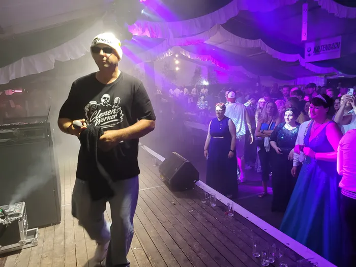 Clemens Werner in schwarzem T-Shirt, weißer Mütze und Sonnenbrille singt auf einer Bühne vor einer Menschenmenge in einem lila beleuchteten Festzelt.