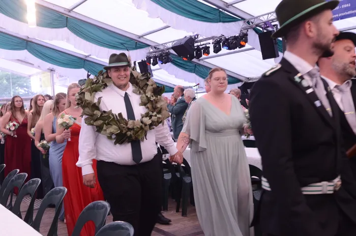Ein Mann mit einem großen Blätterkranz und einer Frau in einem hellen Kleid gehen Hand in Hand durch ein Festzelt.