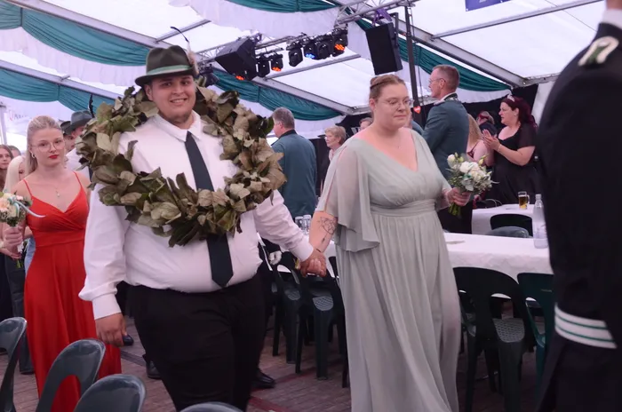 Ein Mann mit einem großen Blattkranz um den Hals und eine Frau in einem hellgrauen Kleid gehen Hand in Hand durch ein Festzelt.