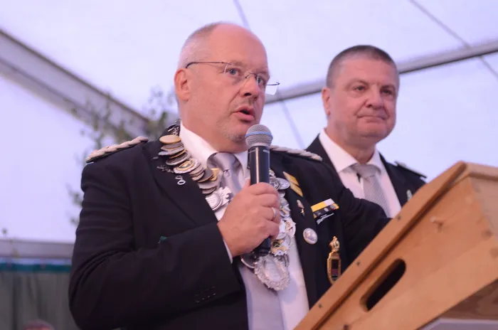 Sascha Kasel spricht in einer schwarzen Uniform mit einer silbernen Königskette an einem Rednerpult in ein Mikrofon.