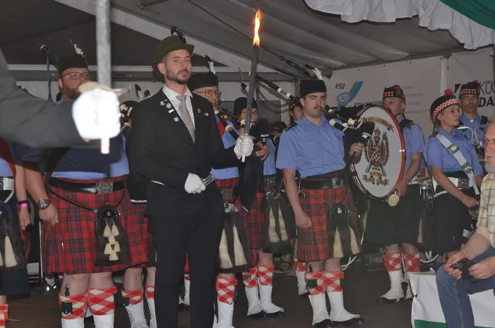 Ein Mann in dunklem Anzug und grünem Hut hält eine brennende Fackel vor einer Gruppe von Männern in Kilts bei der Krönungsfeier des Schützenvereins Eiringhausen.
