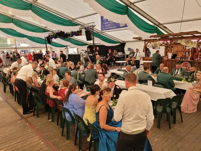 Gäste in festlicher Kleidung und Uniformen sitzen an langen Tischen in einem mit grünen Stoffen dekorierten Festzelt.