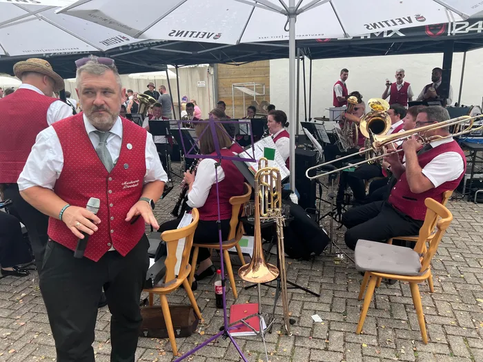 Ein Mann in einer roten Weste spricht in ein Mikrofon, während im Hintergrund eine Blaskapelle unter weißen Sonnenschirmen spielt.