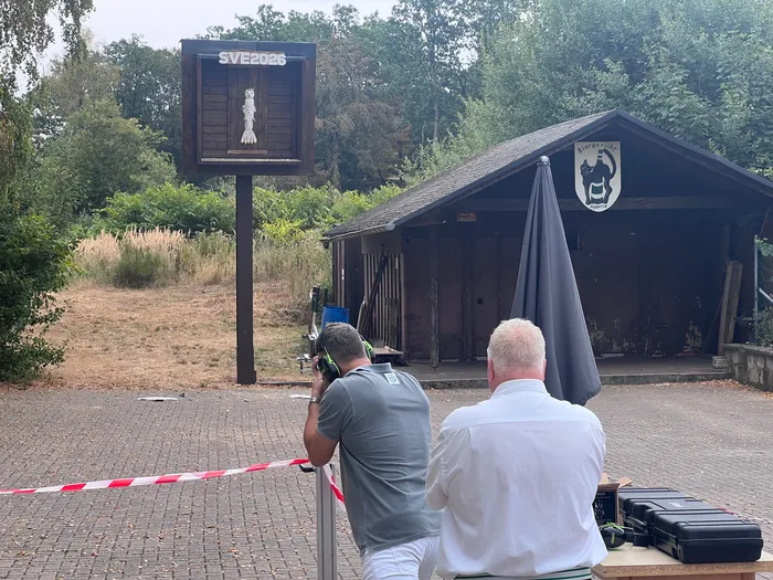 Ein Mann zielt mit einem Gewehr auf einen hölzernen Vogel an einer Zielscheibe mit der Aufschrift „SVE 2026“.