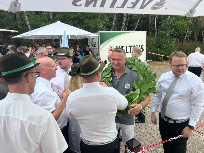 Sascha Kasel trägt einen großen grünen Blattkranz und wird von Männern in weißen Hemden und traditionellen Hüten umringt.