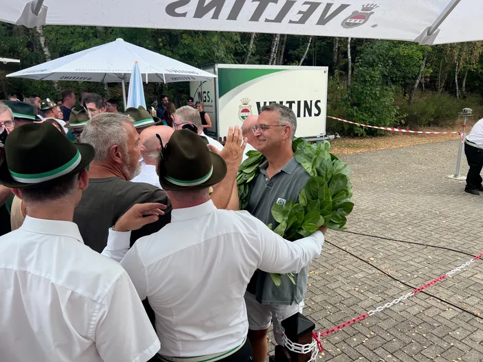 Sascha Kasel trägt einen großen grünen Kranz und wird von einer Gruppe Männer in traditionellen Hüten gratuliert.