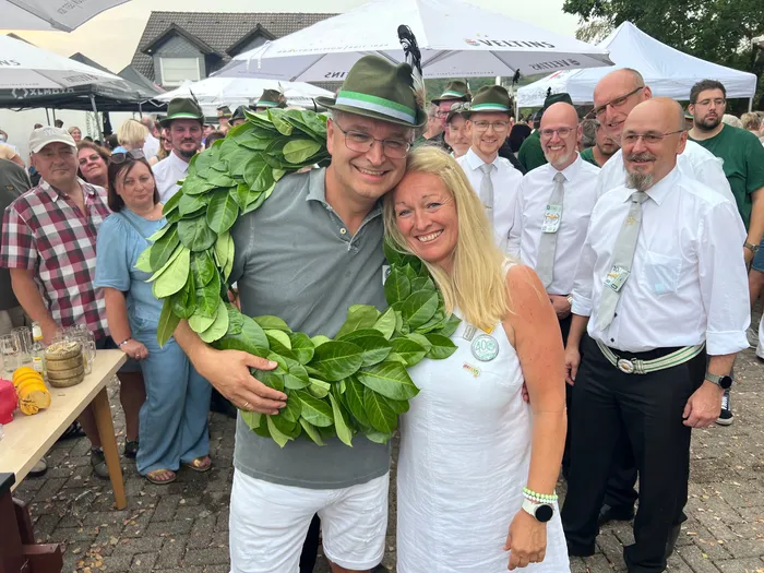 Sascha Kasel trägt einen großen grünen Kranz und einen Hut mit Feder und lächelt gemeinsam mit einer Frau vor einer Gruppe von Festgästen.