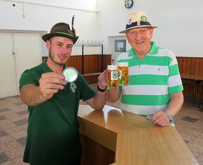 Ein junger Mann hält eine Medaille mit der Zahl 40 hoch, ein älterer Mann in einem grün-weiß gestreiften Poloshirt hält zwei Gläser Bier.