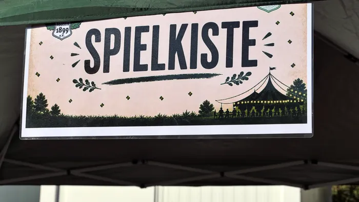 Schild mit der Aufschrift „SPIELKISTE“, dekoriert mit Blättern und der Silhouette eines Festzelts.