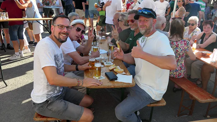 Eine Gruppe von Männern sitzt an Holztischen im Freien, trinkt Bier und macht Handzeichen wie Daumen hoch.
