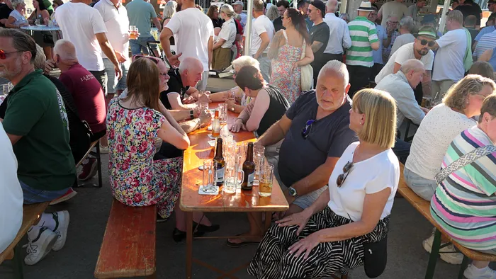 Menschen sitzen an Holztischen im Freien und trinken Bier bei einem Schützenfest-Warm-up.