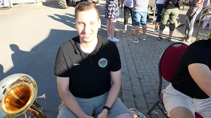 Ein lächelnder Mann in einem schwarzen Poloshirt sitzt neben einem Blechblasinstrument bei einem Schützenfest-Warm-up.