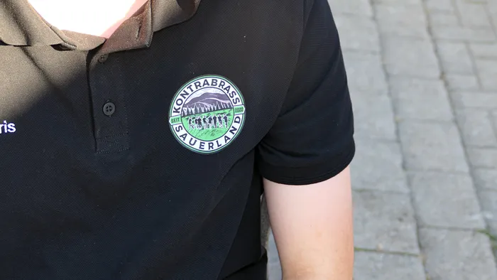 Ein schwarzes Poloshirt mit einem runden, grünen Logo der „Kontra Brass Sauerland“, das eine Berglandschaft und eine Gruppe von Menschen zeigt.