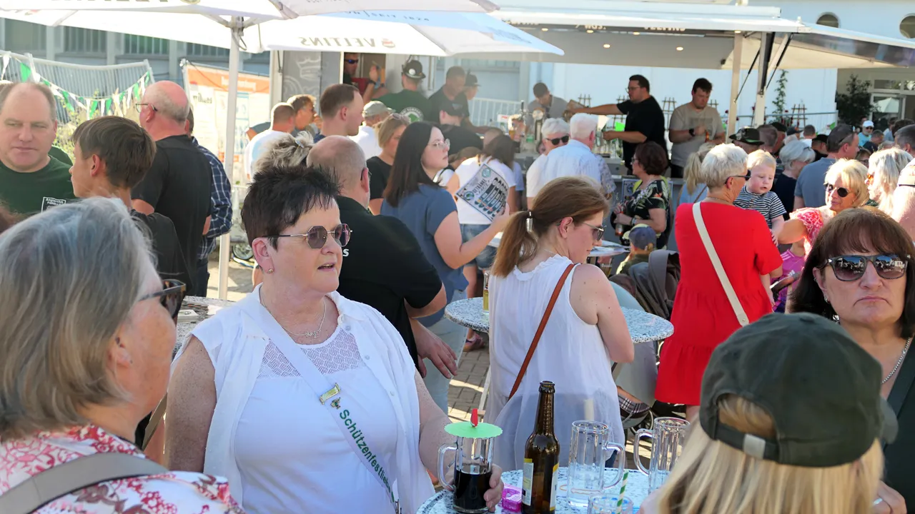 Menschen sitzen an Tischen unter weißen Sonnenschirmen bei einem Schützenfest-Warm-up im Freien.