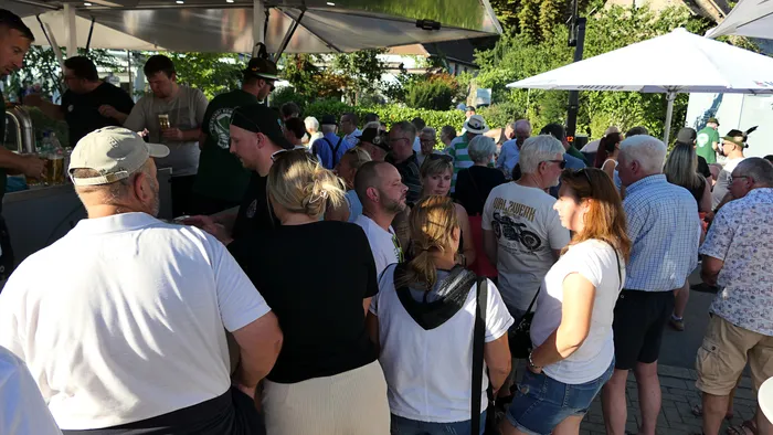 Menschen stehen bei einem Schützenfest-Warm-up unter weißen Sonnenschirmen an einem Tresen im Freien.