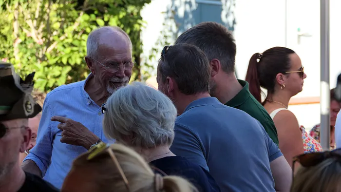 Ein lächelnder älterer Mann in einem hellblauen Hemd zeigt auf eine Gruppe von Menschen bei einem Schützenfest-Warm-up.