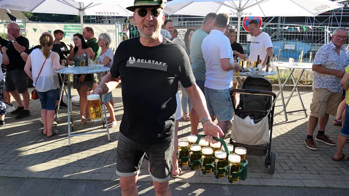 Ein Mann trägt eine Platte mit gefüllten Biergläsern bei einem Schützenfest-Warm-up im Freien unter Sonnenschirmen.