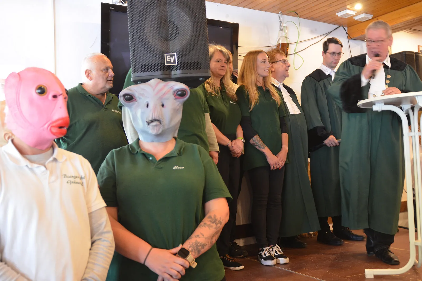 Eine Gruppe von Menschen in grünen Poloshirts und einige in Roben steht vor einem Lautsprecher. Zwei Personen tragen Tierkostüme mit Fisch- und Walgesichtern.