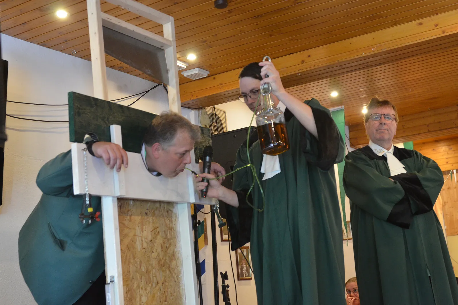 Drei Männer in grünen Roben vollziehen ein Biergericht: Ein Mann schaut durch ein Holzgestell, während ein anderer ihm ein Glas Bier eingießt. Im Hintergrund steht ein weiterer Mann in Robe.