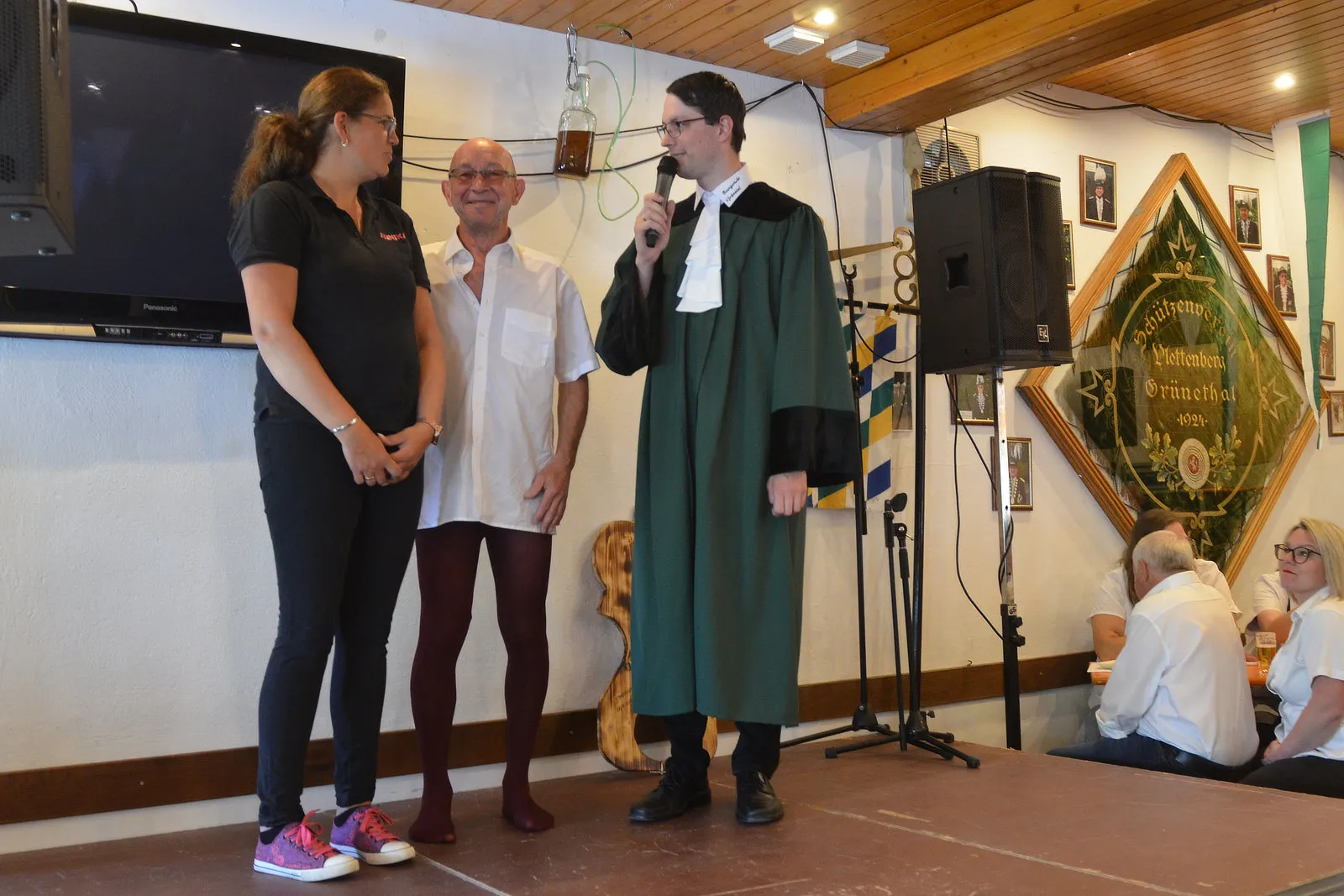 Drei Personen stehen auf einer Bühne vor Publikum: eine Frau in rotem Shirt, ein Mann in weißem Hemd und ein Mann in Robe mit Mikrofon.