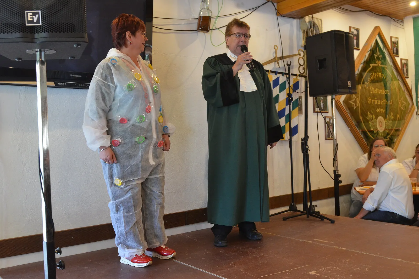 Zwei Personen auf einer Bühne, eine Frau mit roten Haaren und ein Mann in einem dunklen Gewand, sprechen in Mikrofone. Im Hintergrund ein Wappen und Zuschauer an Tischen.