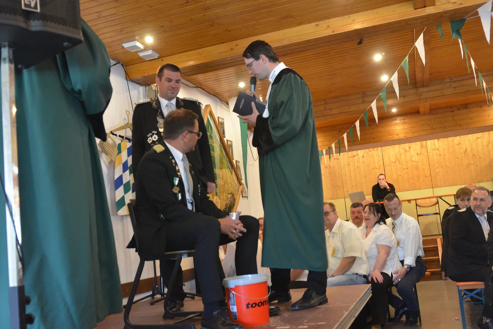 Ein Mann in einem grünen Gewand hält eine Urkunde vor einem sitzenden Mann in Uniform. Im Hintergrund stehen weitere Personen und bayerische Flaggen.