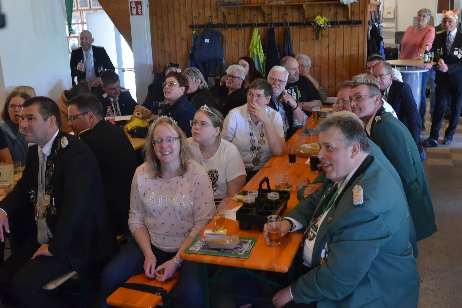 Verschiedene Menschen sitzen an Biertischen in einem Raum mit Holztäfelung und schauen in eine Richtung, einige halten Biergläser. Im Vordergrund eine Frau mit einer Tiara und ein Mann im grünen Trachtenanzug.