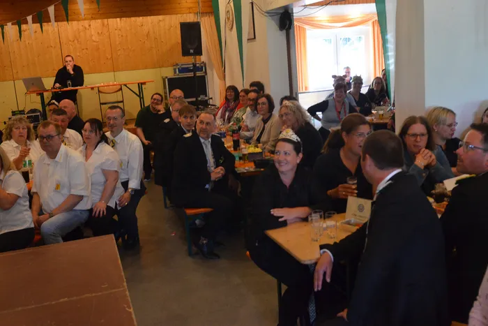 Verschiedene Menschen sitzen an Biertischen in einem hellen Raum und unterhalten sich. Im Hintergrund steht eine Frau an einem Pult und spricht in ein Mikrofon.