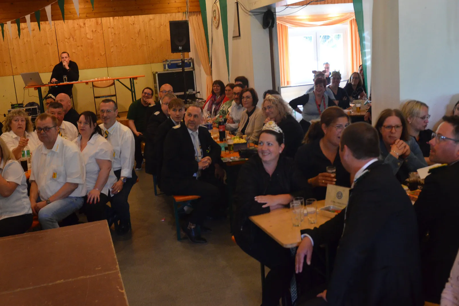 Verschiedene Menschen sitzen an Biertischen in einem hellen Raum und unterhalten sich. Im Hintergrund steht eine Frau an einem Pult und spricht in ein Mikrofon.