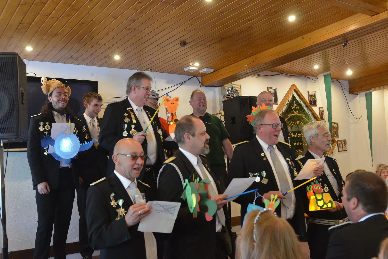 Mehrere Männer in traditionellen Schützenuniformen mit Orden und Hüten stehen in einem Raum mit Holzvertäfelung und singen oder lesen von Blättern. Im Hintergrund ein Wappen und ein Lautsprecher.