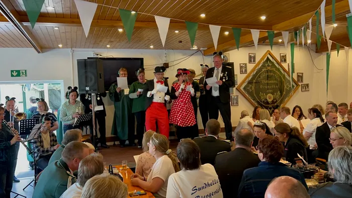 Mehrere Personen stehen auf einer Bühne und singen in ein Mikrofon, während im Hintergrund eine Blaskapelle spielt und Gäste sitzen.