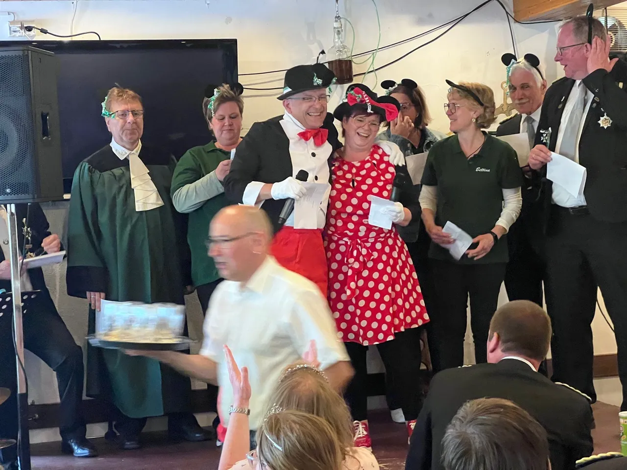 Festlich gekleidete Personen stehen um einen Mann in weißem Hemd, der im Mittelpunkt steht. Im Hintergrund sind weitere Menschen und eine Bühne zu sehen.