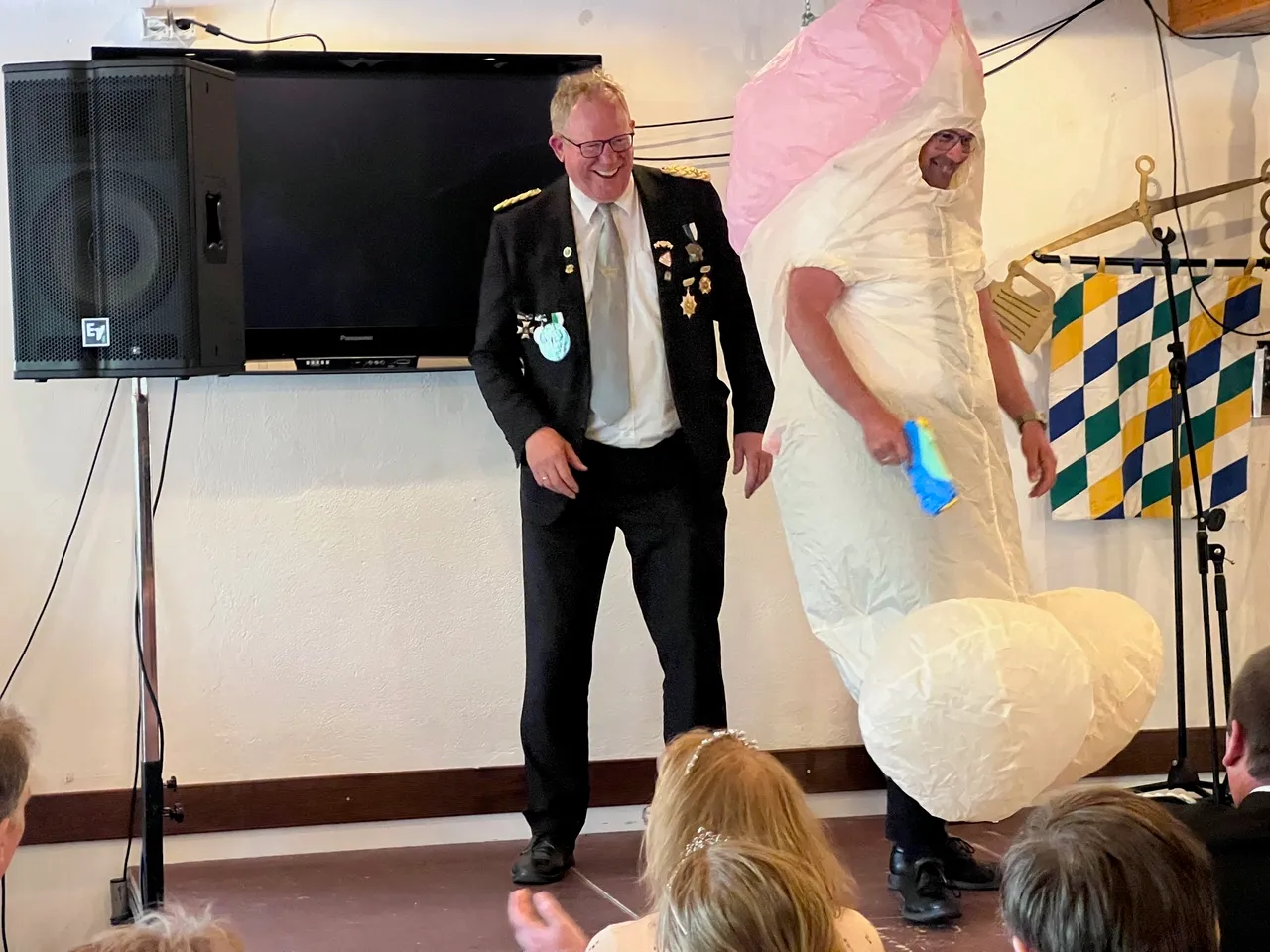 Ein Mann in Uniform lacht und gestikuliert auf einer Bühne, während ein anderer Mann in einem weißen Kostüm mit einer großen rosa Figur interagiert. Im Hintergrund ein Fernseher und ein Banner mit blau-weißen Rauten.