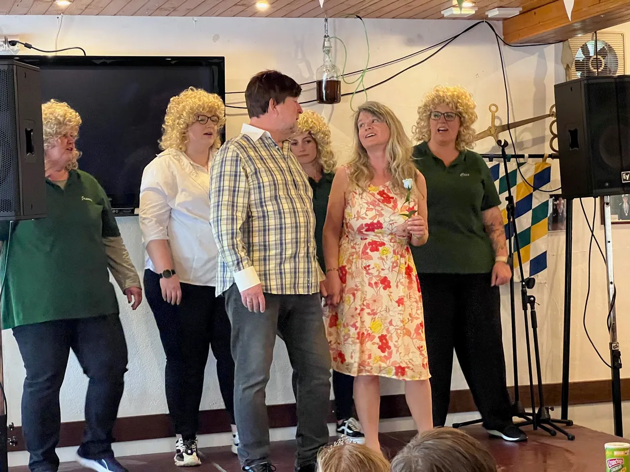 Fünf Personen mit gelockten Perücken stehen auf einer Bühne und singen oder sprechen in ein Mikrofon. Sie tragen grüne Poloshirts und wirken enthusiastisch.