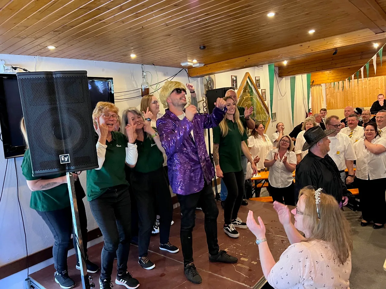 Ein Mann mit blondierten Haaren und Sonnenbrille performt auf einer Bühne vor einem Publikum, das applaudiert. Links stehen Frauen in grünen Oberteilen, rechts ein Mann im schwarzen Anzug und Hut.