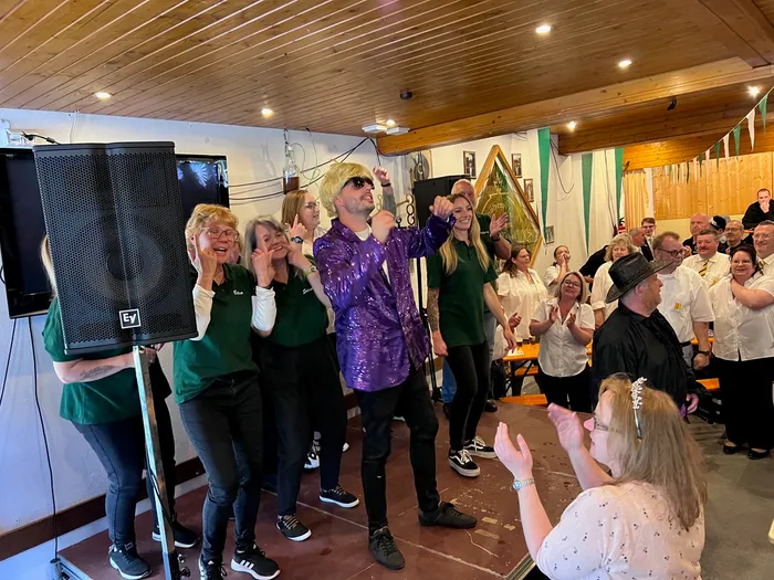 Ein Mann mit blondierten Haaren und Sonnenbrille performt auf einer Bühne vor einem Publikum, das applaudiert. Links stehen Frauen in grünen Oberteilen, rechts ein Mann im schwarzen Anzug und Hut.