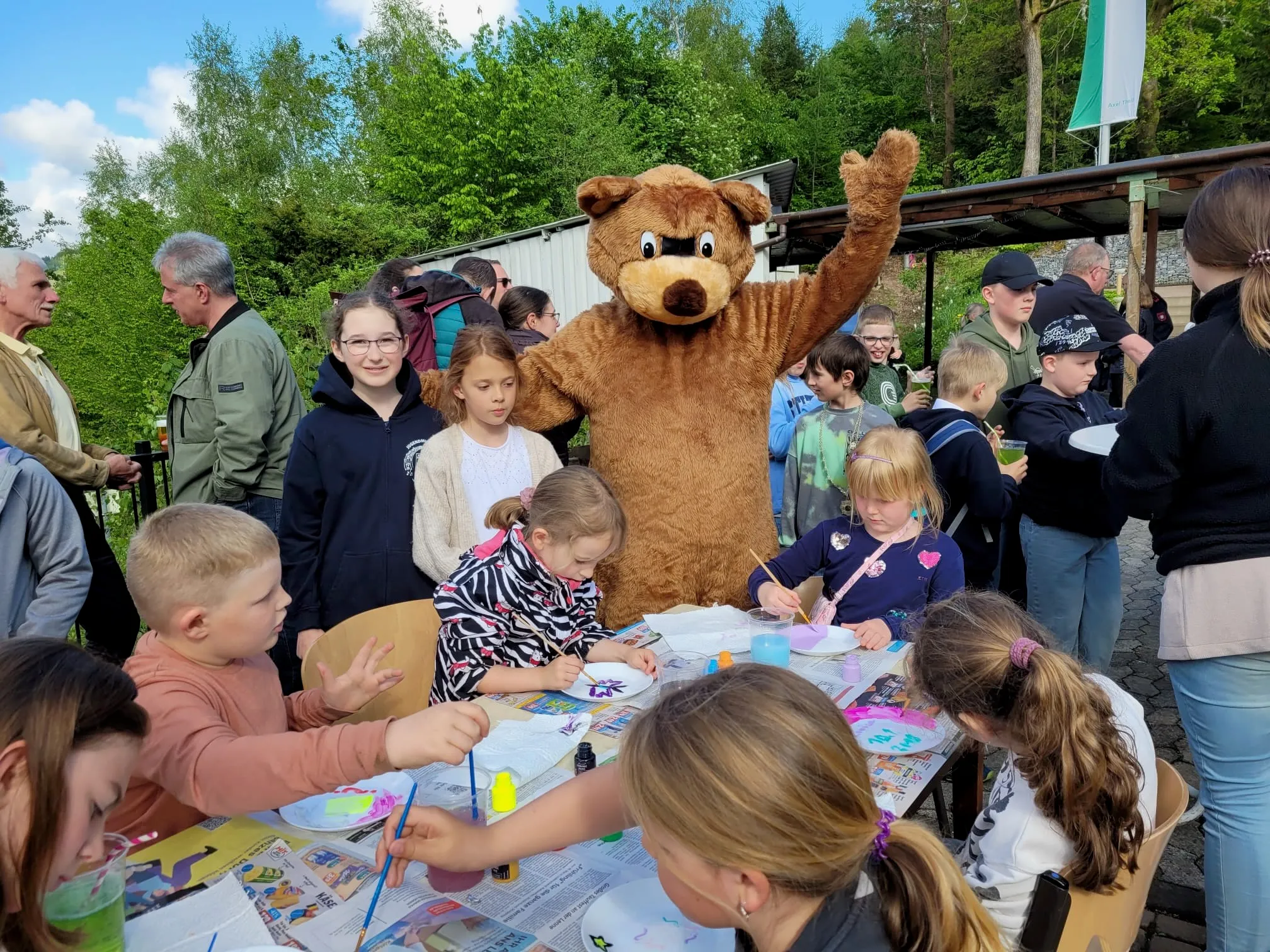 Kinder sitzen an einem Tisch und malen, während ein Bär-Maskottchen im Hintergrund winkt. Im Hintergrund stehen weitere Menschen und beobachten die Aktivitäten.