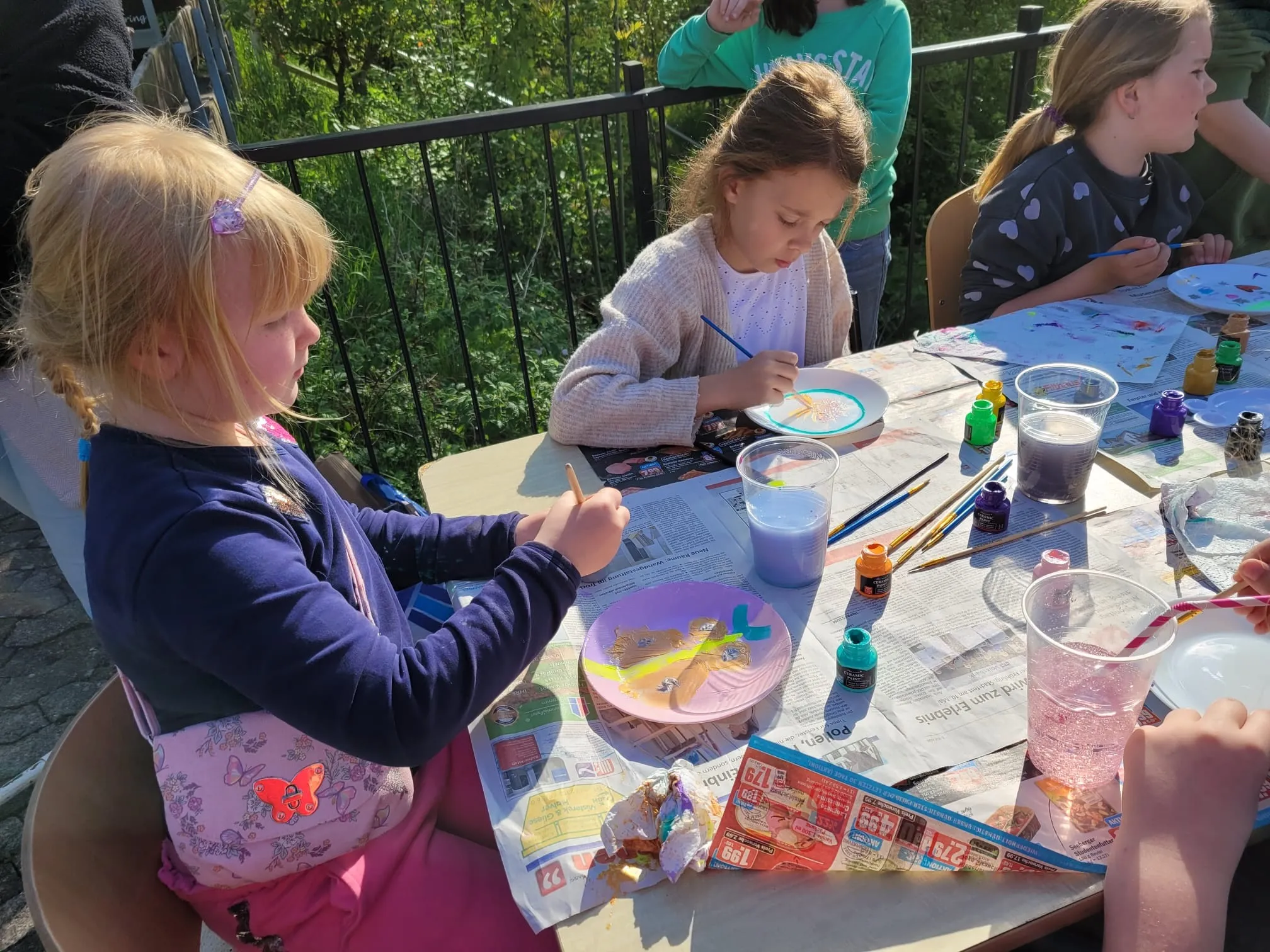 Mehrere Kinder sitzen an einem Tisch im Freien und bemalen Teller mit bunten Farben. Im Hintergrund sind weitere Kinder und ein rotes Gebäude zu sehen.