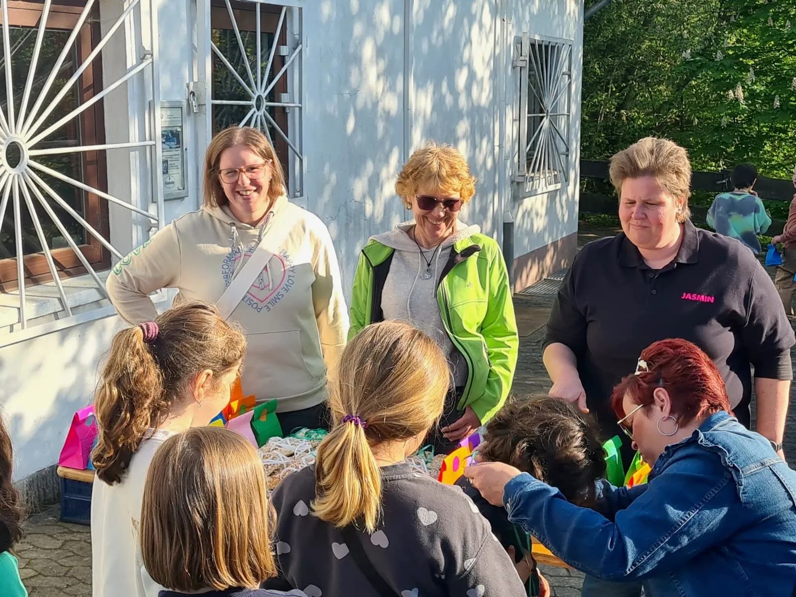 Mehrere Frauen und Kinder sitzen oder stehen auf einem gepflasterten Platz vor einem Gebäude mit großen Fenstern und einem dekorativen, sonnenförmigen Element an der Fassade. Sie scheinen sich bei einer Veranstaltung im Freien zu unterhalten und basteln.
