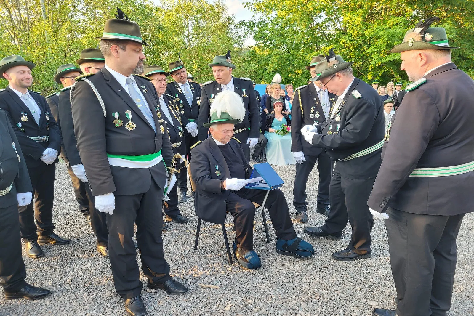 Bei der Krönung des Schützenkönigs überreicht ein Mann in Uniform einem sitzenden Mann eine Urkunde. Um sie herum stehen weitere Mitglieder des Schützenvereins in grünen Uniformen mit weißen Handschuhen und Dekorationen.
